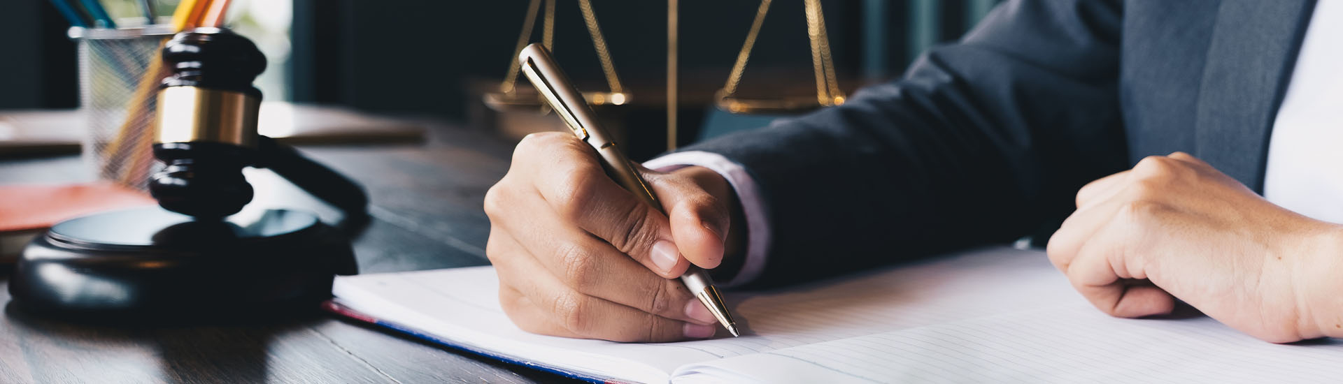 Mano escribiendo en un documento con balanza y mazo de justicia al fondo